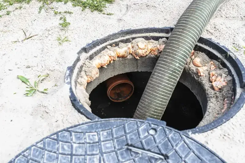 Vacuum hose lowered into an open sewer or septic access hole with the cover set beside it